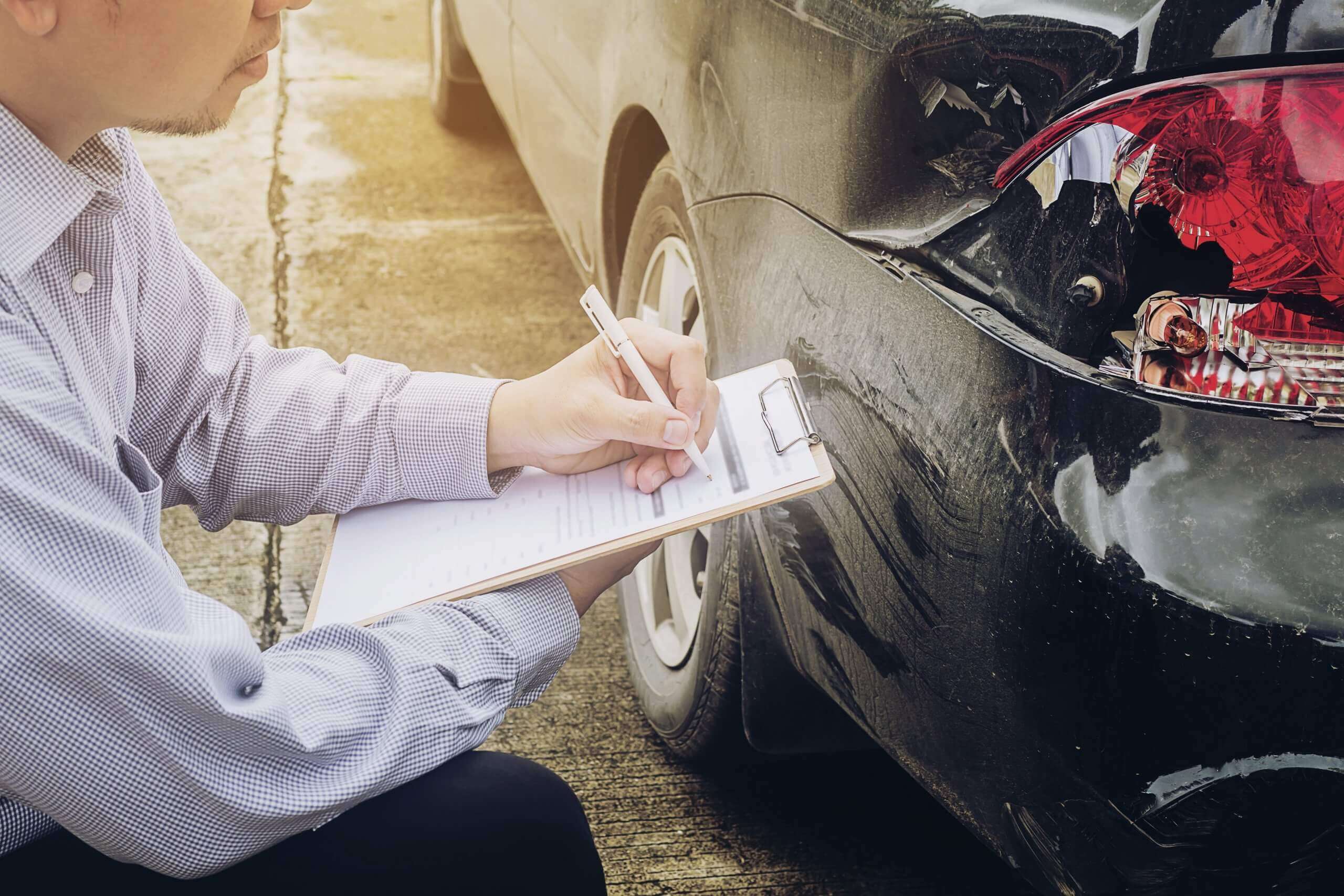 Ein Mann mit einem Klemmbrett begutachtet den Unfallschaden an einem Auto, nachdem die Versicherung die Kosten nicht übernommen hat